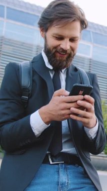 Siyah ceketli, bakımlı sakallı ve kotlu güzel görünümlü genç ofis çalışanının portresi. Mobile 'deki eğlenceli resimlerden gülerken büyük şehir binasının arka planında durmuş.