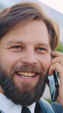 Güzel görünümlü, neşeli, 30 yaşında, sakallı ofis çalışanı. Akıllı telefondan konuşan, interlocutors şakalarından gülümseyen, modern, güzel şehir binasının yanında duran.