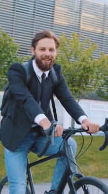 Güzel görünümlü, neşeli, mutlu, 30 yaşında, siyah ceketli ve kot pantolonlu, etkileyici modern bina arka planında bisikletle poz veren erkek ofis müdürü.