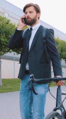 Sevimli dengeli başarılı 30 yaşında, sakallı, siyah ceketli ve kot pantolonlu bir ofis çalışanı geniş şehir yolunda bisikletle yürüyor ve mobil sohbetler yapıyor, yavaş çekim.