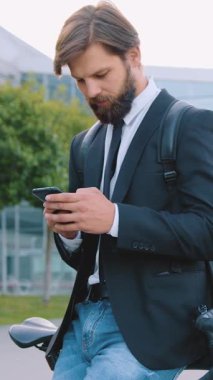 İyi huylu, 30 yaşında, bakımlı, siyah ceketli ve kot pantolonlu bir adam kaldırımda akıllı telefon kullanırken bisikletine yaslanıyor.