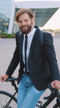 İyi huylu, neşeli, kendine güvenen koyu renk ceketli ve kot pantolonlu bir ofis çalışanı. Kombine yolda bisikletinin yanında duruyor ve şanslı bir gülümsemeyle kameraya bakıyor.