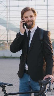 Modern şehir panoramik binasının yakınında bisikletinin üzerinde dururken akıllı telefonuyla konuşan hoş, mutlu, başarılı, genç iş adamının portresi.