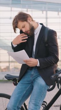 Güzel görünümlü, ciddi bir iş adamı 30 yaşında, resmi giysiler içinde, modern şehir panoramik inşaatı sırasında iş arkadaşıyla telefon görüşmeleri yaparken bisikletine yaslanıp finansal belgeleri tartışmakla meşgul.