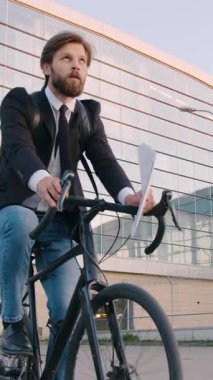 İyi sonuç veren iş konsepti, güzel görünümlü başarılı 30 yaşında ofis yöneticisi sakallarını etrafa saçarken panoramik pencereli büyük şehir binasının yanında bisiklet sürüyor.