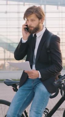 Yakışıklı, yoğun sakallı, resmi giysiler içinde, bisikletine yaslanmış, cep telefonuyla konuşan ve panoramik pencereli modern şehir inşaatı yakınlarındaki finansal belgeleri okuyan genç bir işadamı.