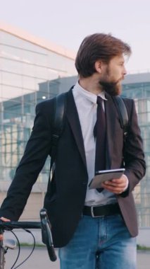 Güzel görünüşlü, 30 'lu yaşlarda, iş kıyafetleri içinde sakallı, elinde tablet tutarken büyük, modern panoramik yapı arka planında kendi bisikletiyle gezen iyi görünümlü, odaklanmış bir adam.