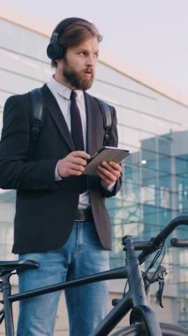 Yakışıklı, kendine güveni tam, 30 yaşında, resmi kıyafetleri ve kulaklıkları olan, camlı şehir binasının yakınındaki i-pad 'de çalışan ofis müdürünün ön görüntüsü.