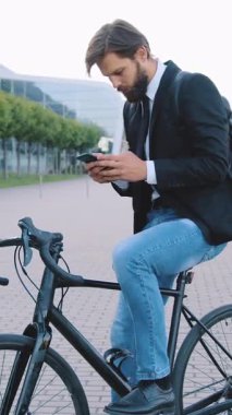 Güzel görünümlü, 25-30 yaşlarında, koyu renk ceketli, kot pantolonlu, sakallı, erkek ofis müdürü şehir binasının yanındaki patikada bisikletinin üstünde duruyor.