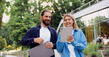 Tablet ve iş ortağını elinde tutan genç bir kadın birlikte ekrana bakıyor, açık hava iş görüşmeleri sırasında çalışma süreçleri üzerine odaklanmış bir tartışmaya girişiyor.
