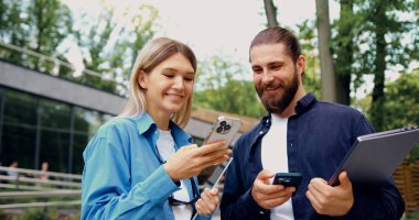 Akıllı telefon ve tablet tutan kadınla akıllı telefon ve dizüstü bilgisayar tutan adamla işbirliği yapan erkek ve kadının portresi. Telefon ekranında anahtar verileri gösteriyor. Hem gözden geçiriyor hem de gülümsüyor.