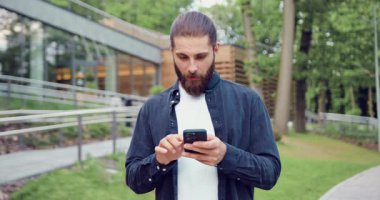 Şehir parkında akıllı telefon kullanan ve beklenmedik haberlere duygusal tepki veren beyaz bir adam. Çevrimiçi iletişim ve teknoloji kavramı. Yavaş çekim