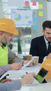 Açık yeşil yelek giyen, kendine güvenen, sakallı, tecrübeli bir mühendis, ofis odasındaki diğer müteahhitlerle yaptığı ortak toplantıda başarılı, sakallı bir yöneticiyle inşaat projesinin detaylarını tartışıyor.