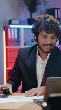 Yakışıklı gülümseyen başarılı kıvırcık sakallı müslüman ofis yöneticisi, kablosuz kulaklıklarla modern çalışma odasındaki bilgisayarda karanlık zamanlarda toplantı düzenliyor.