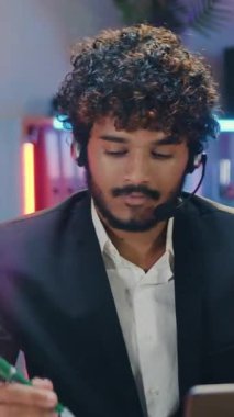 Sempatik gülümsemeli, başarılı kıvırcık Arap iş adamı resmi kıyafetli, iş arkadaşlarıyla çalışma odasında iş görüşmeleri, gece aydınlatmaları ve iş konsepti olan bilgisayarla konuşurken kulaklık takıyor.