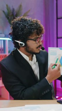 İşletme konsepti. Yüksek yetenekli, kıvırcık sakallı, kablosuz kulaklıklı Müslüman ofis yöneticisi geceleri kameraya konuşuyor. Çalışma odasını aydınlatıyor ve izleyiciler için bilgi sunuyor.