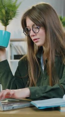 Ev eğitimi konsepti, oldukça yetenekli 25 yaşında, uzun kahverengi saçlı bir kızın rahat ev ofisinde masada oturup aynı anda dizüstü bilgisayarında çayını yudumlarken...