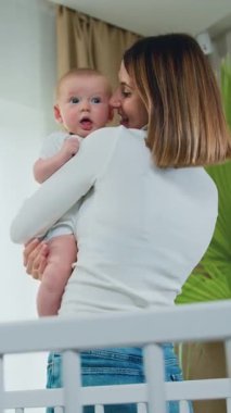 Sevgi dolu genç bir anne, rahat bir bebek odasında durup sarılır ve güzel bebeğini beyaz bir elbise içinde sakinleştirir. Çocukluk kavramı, yeni hayat ve ebeveynlik. Yavaş çekim