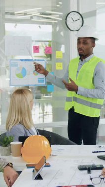 İş arkadaşları ve yöneticileriyle ortak inşaat projesinin detaylarını tartışan yelek ve başlık takan iyi görünümlü, siyah derili inşaat firması uzmanı.