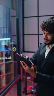Yakışıklı, motive edici, siyah giyimli, kıvırcık sakallı Arap iş adamı tablet cihazıyla ilgili bilgi okuyor ve çalışma odasındaki işaretle şeffaf tahtada kayıt yapıyor.