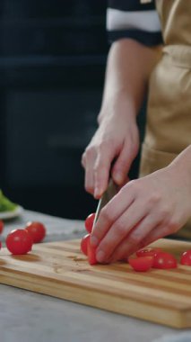 Önlüklü tanınmayan bir kadın besleyici vitamin salatası hazırlarken kesme tahtasındaki keskin bıçakla yarım kiraz domateslerini kesiyor.