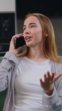 Telefonla konuşan gülümseyen genç bir kadın evdeki kanepede oturuyor. Modern koltukta cep telefonuyla flört eden güzel bir kadının portresi. Gülümseyen kız mutfakta sohbet ediyor. Yavaş çekim,