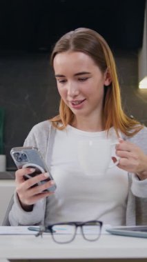 Güzel sarışın bir kadının portresi akıllı telefon kullanarak sosyal medyada sabahları evde yalnız başına kahve içiyor. Evde cep telefonuyla konuşan bir kadın var. Yavaş çekim.