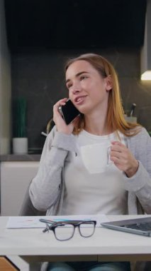 Genç, mutlu bir kadın elinde kahve fincanıyla akıllı telefonuyla bir telefon görüşmesi yapıyor. Modern mutfakta gülümseyen serbest çalışan kadın telefonda uzaktan konuşuyor. Yavaş çekim.