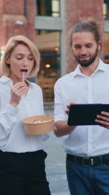 Bearded smiling man shows something on a tablet to a woman. Blonde woman is eating a salad and talking to a man while walking down the street. Slow motion.