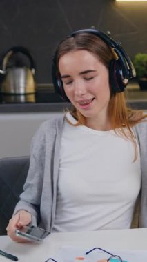 Mutlu, rahat kadın, dizüstü bilgisayar başında oturmuş kulaklık takarak evde modern mutfakta akıllı telefon kullanarak müzik dinliyor. Yavaş çekim.