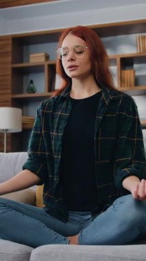 Pratik, iç ahengi ve dengeyi hisseder. Stres yok. Çekici kızıl saçlı beyaz kadın Lotus pozisyonunda yoga yapıyor ve meditasyonun gözlerini kapıyor. Evdeki koltukta oturuyor. Yavaş çekim.