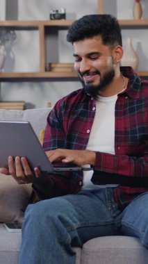 Genç, güzel, yetişkin Hintli bir adam rahat bir oturma odasında elinde dizüstü bilgisayarla oturuyor. Yaratıcı İspanyol erkek sosyal medyayı kontrol ediyor, internette geziniyor. Yavaş çekim.