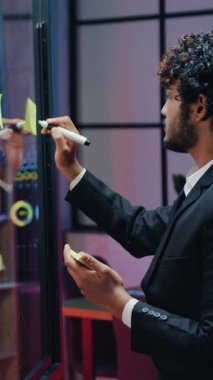 Sorumluluk sahibi, başarılı, başarılı, yüksek vasıflı, kıvırcık saçlı, şeffaf bir ofis çalışanı geceleri çalışma odasını aydınlatır ve yapışkan notlarla kayıt yapar ve bu tahtaya yapıştırır.