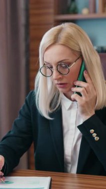 İş konsepti, resmi giyinmiş gözlüklü, deneyimli yetişkin bir iş kadınının bilgisayardaki çalışmalarını finansal raporlar ve mobil konuşmalarla birleştirmesi.