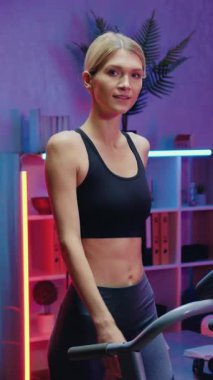 Güzel görünüşlü, formda, spor elbiseli, spor elbiseli, yürüyüş egzersizleri yapan ve gece yarısı evin arka planındaki kameraya bakan sarışın.