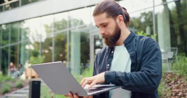 Modern bir ofis binasının yakınında kablosuz bir laptop kullanan yakışıklı girişimci, çevrimiçi iş ve uzaktan çalışmayı yönetirken daktilo ve kaydırma yapıyor. İş, teknoloji ve dijital yaşam tarzı kavramı. Yavaş çekim