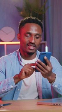 Sevimli, mutlu, rahat, Afro-Amerikalı gencin portresi. Rahat ev ofisinde bilgisayardaki fazla mesai sırasında cep telefonundaki arkadaşlarıyla mesajlaştığı mesajları cevaplıyor.