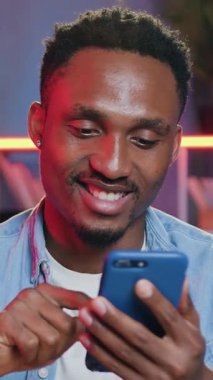 25-30 yaşlarında, renkli gece lambalarıyla kameranın önünde oturan ve akıllı telefon kullanan, siyah tenli, yakışıklı, gülümseyen bir adam.