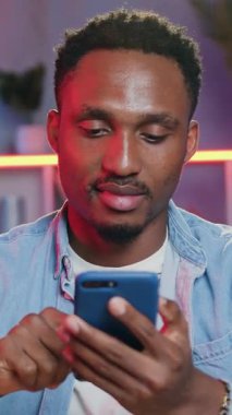 Sevimli, rahat, 30 yaşında Afro-Amerikan haberleri sosyal medyada akıllı telefondan okunuyor ve renkli ışık efektleri olan güzel dekore edilmiş bir odada kameraya bakıyor.