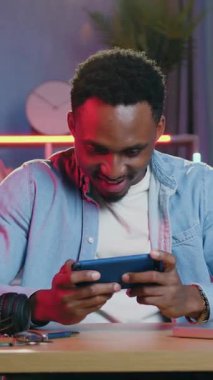 Sevimli, mutlu, 30 yaşında, siyah tenli bir adamın portresi. Akıllı telefondan oyun oynayan ve kazanırken eğlenen ve renkli ışık efektleri, eğlence ve başarı konseptiyle sıcak bir odada el kaldıran.