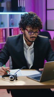 Sevimli bir gülümseme başarılı, kıvırcık sakallı Arap ofis müdürü resmi giyim ve gözlükleriyle modern çalışma odasında gece ışıklarıyla bilgisayarda yazı yazıyor.