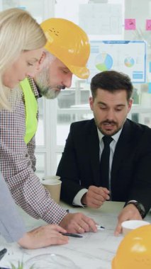 Toplantı odasında yapılan brifingde ciddi derecede deneyimli sakallı ana yöneticiyle birlikte yeni binanın inşaat taslağını analiz eden yelekli, çok ırklı ve çalışkan mimarların portresi.