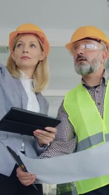 Yakışıklı, 50 yaşında, miğfer ve yelek giyen, yakışıklı, kendine güvenen kadın-müşterisiyle birlikte inşaat alanında yürüyen ve ortak inşaat projesinden bahseden,