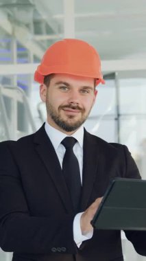 İyi görünümlü, modern, deneyimli, sakallı bir mimar veya güvenlik kaskı takmış bir mühendis. Tablet cihazıyla çalışıyor, sonra da kapalı alandaki inşaat alanındaki kameraya bakıyor.