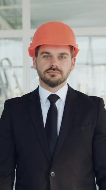 İyi görünümlü, yüksek vasıflı, şık sakallı, takım elbiseli ve kasklı bir mimar. Kameraya çapraz kollu bakıyor, inşaat alanında duruyor.