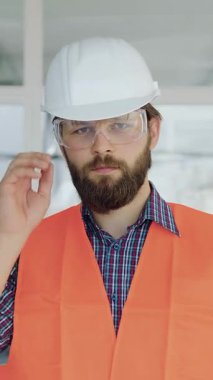 35 yaşında, güvenlik kaskı, gözlük ve yelek takan firma sakallı çekici bir yönetici. Bitmemiş ofiste kameraya poz veriyor.