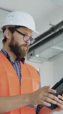 Çekici, yüksek yetenekli, maksatlı, sakallı inşaat firması, koruyucu gözlük ve yelek uzmanı portresi. Tablet cihazında inşaat planının detaylarını kontrol ediyor, tamamlanmamış bir binanın içinde duruyor.