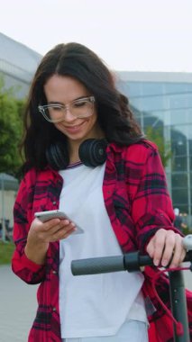 Mode gözlüklü, şık, neşeli esmer kadın geniş caddede kişisel elektrikli scooterıyla geziyor. Şehir binasının arka planında ve cep telefonunu kullanıyor. Kapatın.