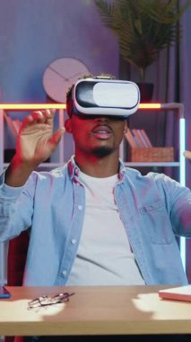 Modern teknoloji konsepti, 30 yaşında, kendine güveni tam bir Afro-Amerikan erkeği rahat bir odada gece ışıklarıyla oturuyor ve sanal gerçeklik kulaklığıyla hayali ekran üzerinde çalışıyor.