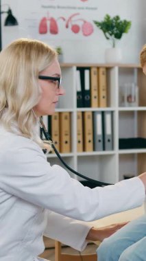 Hoş, sakin, 12 yaşında bir okul çocuğu rahat bir sağlık dolabında masanın üzerinde oturmuş, oldukça yetenekli, açık renk saçlı, dost canlısı, ciğerlerini dinleyen bir bayan çocuk doktoru.